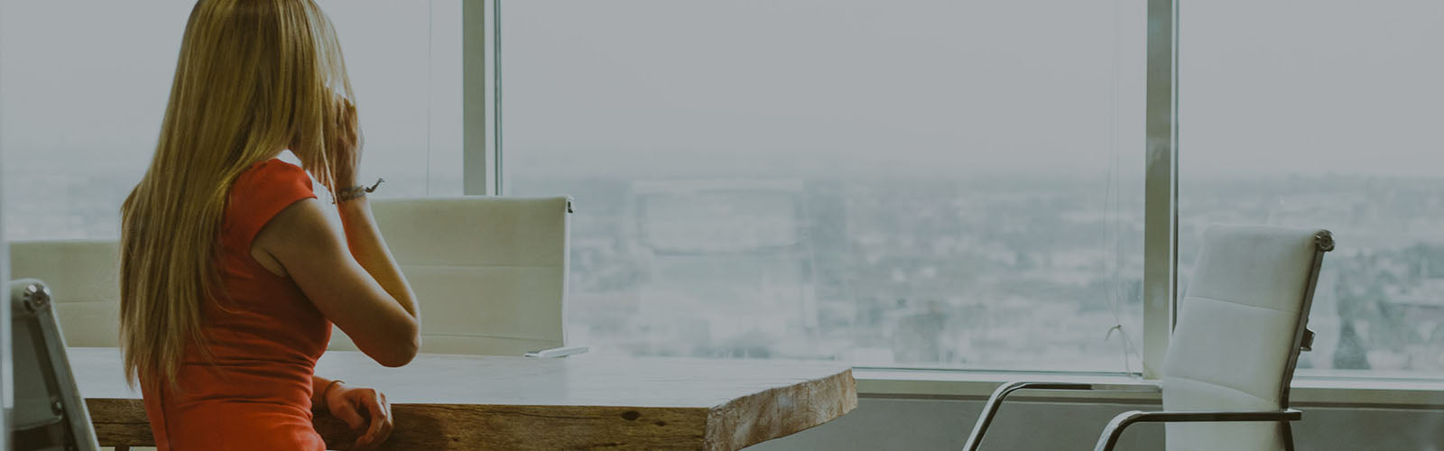Eine blonde Frau in rotem Kleid sitzt an einem Besprechungstisch in einem Büro mit Blick über die Stadt und telefoniert